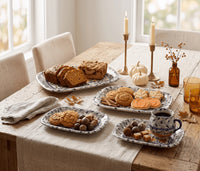 A styled dining table scene with multiple ceramic serving trays holding sliced bread, cookies, acorns, and a cup of coffee. The setting includes chairs, candles, pumpkins, leaves, and amber glassware in a bright indoor room.
