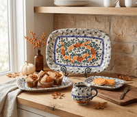 A ceramic serving set displayed on a wooden kitchen counter, including a large rectangular platter, a matching mug, and a small plate, all decorated with a blue and orange floral wreath pattern on a white background. The scene is styled with autumn props like pumpkins, fall leaves, and baked goods.