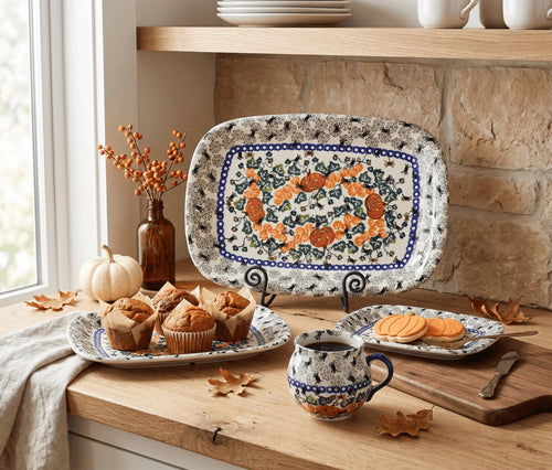A ceramic serving set displayed on a wooden kitchen counter, including a large rectangular platter, a matching mug, and a small plate, all decorated with a blue and orange floral wreath pattern on a white background. The scene is styled with autumn props like pumpkins, fall leaves, and baked goods.