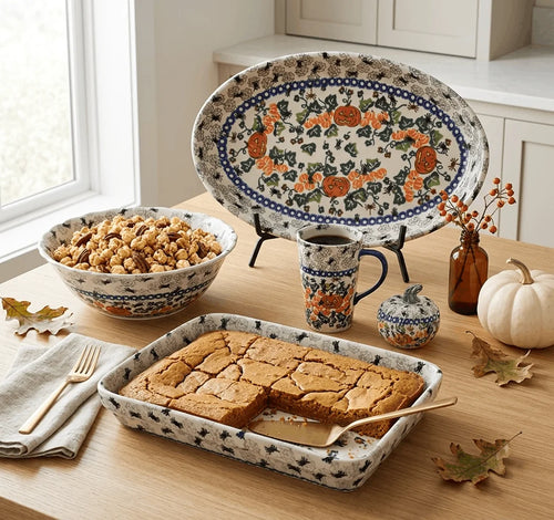A set of ceramic autumn-themed tableware from Polish Pottery, including an oval platter, a bowl, a mug, a small lidded pot, and a rectangular baking pan, styled on a wooden table with fall leaves, a pumpkin, and food.