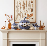 A styled mantel display featuring three blue and white speckled ceramic pumpkins with carved jack-o'-lantern faces, alongside a matching blue and white ghost figurine, a brown glass bottle with orange berries, small white pumpkins, and brass candlesticks, set against a white wall with a botanical art print hanging above.