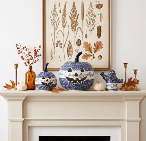 A styled mantel display featuring three blue and white speckled ceramic pumpkins with carved jack-o'-lantern faces, alongside a matching blue and white ghost figurine, a brown glass bottle with orange berries, small white pumpkins, and brass candlesticks, set against a white wall with a botanical art print hanging above.