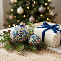 Two decorative Christmas ornaments with a floral and bird pattern in blue, red, and green on a cream background, styled on pine branches next to a wrapped gift box with a blue ribbon, with a blurred Christmas tree in the background.