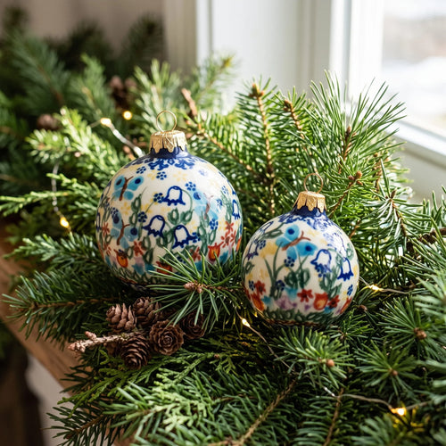 Two hand-painted ceramic Christmas ornaments with a blue and white floral pattern, nestled in green pine branches with small pinecones and warm string lights.