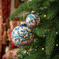 Two hand-painted ceramic Christmas ornaments with floral patterns hanging on a green Christmas tree branch with warm string lights.