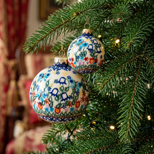 Two hand-painted ceramic Christmas ornaments with floral patterns hanging on a green Christmas tree branch with warm string lights.