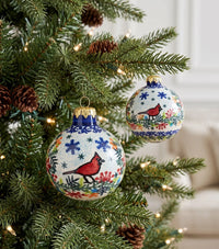 Two hand-painted Christmas tree ornaments with red cardinals and blue floral patterns hanging on a decorated Christmas tree.