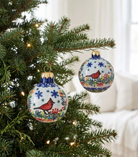 Two hand-painted ceramic Christmas ornaments hanging on a decorated evergreen tree. The ornaments are white with blue floral patterns, red cardinals, and gold caps, set against a softly blurred white living room background.