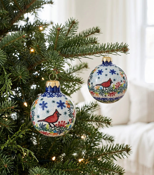 Two hand-painted ceramic Christmas ornaments hanging on a decorated evergreen tree. The ornaments are white with blue floral patterns, red cardinals, and gold caps, set against a softly blurred white living room background.