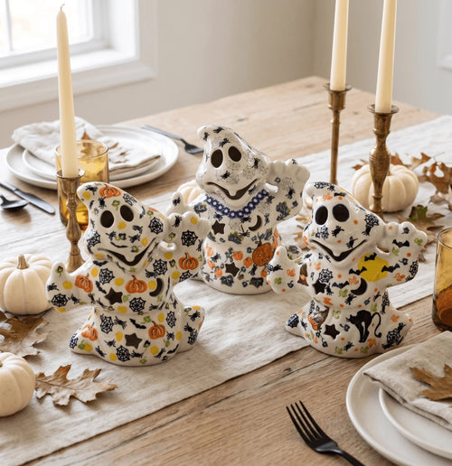Three ceramic Halloween ghost figurines decorated with black, orange, and yellow spooky patterns, arranged on a rustic wooden dining table with fall table settings, candles, and pumpkins.