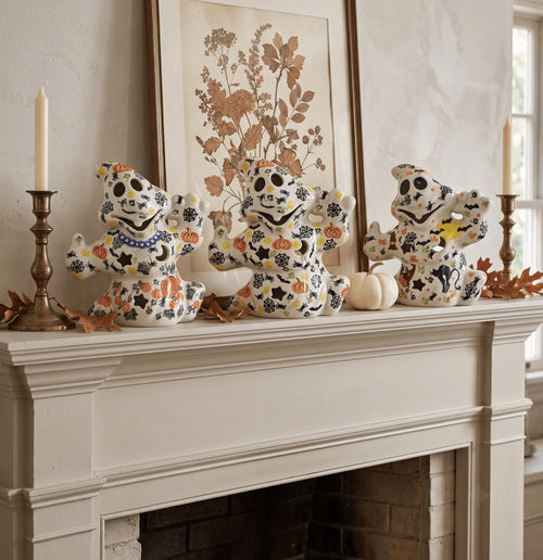 Three decorative ceramic sugar skull figurines with orange and blue floral patterns are arranged on a white fireplace mantel alongside brass candlesticks, a white candle, small pumpkins, and autumn leaves. A framed botanical print hangs on the wall behind them.