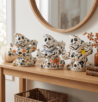 Three ceramic Halloween ghost figurines decorated with orange pumpkins, black bats, and yellow stars, displayed on a wooden console table in a home setting.