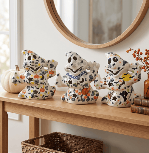 Three ceramic Halloween ghost figurines decorated with orange pumpkins, black bats, and yellow stars, displayed on a wooden console table in a home setting.