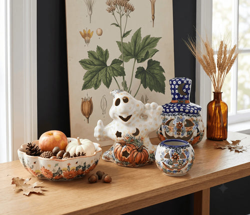 A styled tabletop display featuring a white ceramic ghost figurine, a white ceramic pumpkin-shaped container, a blue-and-white patterned lidded jar, a matching small bowl, and an amber glass vase with dried wheat, all arranged on a wooden table in front of botanical wall art.