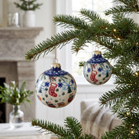 Two round Christmas tree ornaments hanging from pine branches in a cozy indoor setting. The ornaments are cream-colored with blue borders, decorated with red stockings, blue snowflakes, and small holiday motifs.