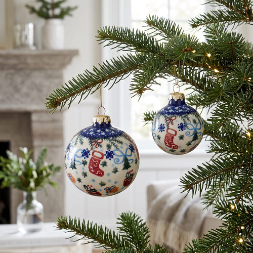 Two round Christmas tree ornaments hanging from pine branches in a cozy indoor setting. The ornaments are cream-colored with blue borders, decorated with red stockings, blue snowflakes, and small holiday motifs.