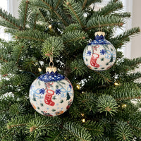 Two round Christmas tree ornaments hanging on a green evergreen tree with warm string lights. The ornaments are white with blue snowflake patterns, red stockings, and small orange and blue decorative details, with gold caps and hangers.