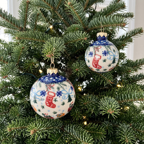 Two round Christmas tree ornaments hanging on a green evergreen tree with warm string lights. The ornaments are white with blue snowflake patterns, red stockings, and small orange and blue decorative details, with gold caps and hangers.