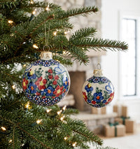 Two hand-painted ceramic Christmas ornaments with floral patterns hang on a decorated evergreen tree. The larger ornament features blue, red, and yellow flowers on a white background, while the smaller one is partially visible behind it.