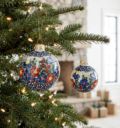 Two hand-painted ceramic Christmas ornaments with floral patterns hang on a decorated evergreen tree. The larger ornament features blue, red, and yellow flowers on a white background, while the smaller one is partially visible behind it.