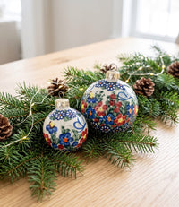 Two hand-painted ceramic Christmas ornaments with floral patterns in blue, red, and yellow on a white background, displayed on a wooden table with pine branches, pinecones, and fairy lights.