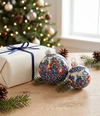 Two hand-painted Christmas ornaments with floral patterns in red, blue, green, and white, displayed on a wooden table next to a wrapped gift box with a blue ribbon, with a blurred Christmas tree in the background.