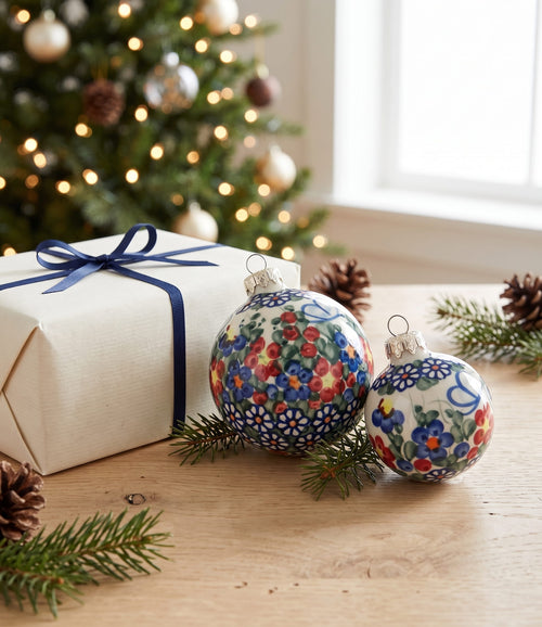 Two hand-painted Christmas ornaments with floral patterns in red, blue, green, and white, displayed on a wooden table next to a wrapped gift box with a blue ribbon, with a blurred Christmas tree in the background.