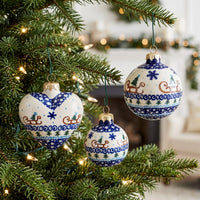 Three ceramic Christmas ornaments with blue and white holiday patterns hang on a green Christmas tree with warm string lights. The ornaments feature designs including sledding, snowflakes, pine trees, and blue wave borders.