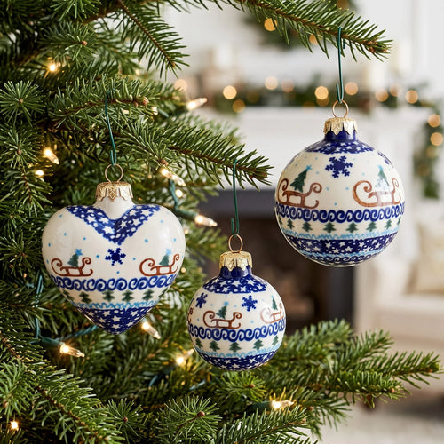 Three ceramic Christmas ornaments with blue and white holiday patterns hang on a green Christmas tree with warm string lights. The ornaments feature designs including sledding, snowflakes, pine trees, and blue wave borders.
