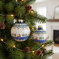 Two round ceramic Christmas tree ornaments with blue, green, and red holiday patterns hang on a pine tree decorated with lights and pinecones in a softly blurred indoor setting.