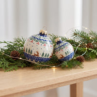 Two decorative Christmas ornaments with blue and white winter landscapes, displayed on a wooden table with pine branches, pinecones, and warm fairy lights.
