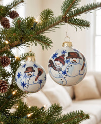 Two round white Christmas ornaments with hand-painted snowmen hanging from a decorated evergreen tree branch in a softly lit indoor setting.