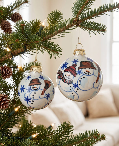 Two round white Christmas ornaments with hand-painted snowmen hanging from a decorated evergreen tree branch in a softly lit indoor setting.
