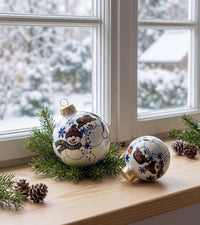 Two decorative Christmas ornaments with painted snowman designs rest on a wooden windowsill beside green pine branches and pinecones, with a snowy outdoor scene visible in the background.